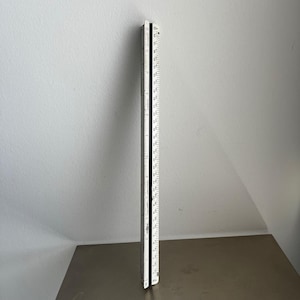 May include: A white and black folding ruler, standing upright. The ruler is marked with inch measurements. The ruler is made of wood and has a black hinge. The background is a light gray wall and a brown surface.