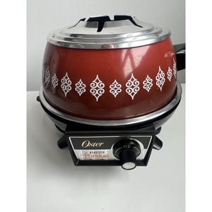 May include: A vintage Oster electric fondue pot. The pot is a deep red with white decorative patterns. It has a silver-colored lid and a black base with a control panel that reads "Oster electric fondue".