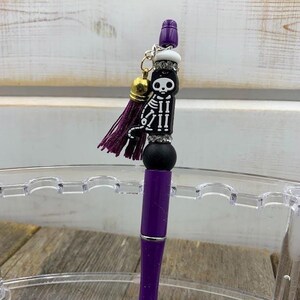 May include: Purple pen with a black and white skeleton cat charm with rhinestones and a purple tassel.