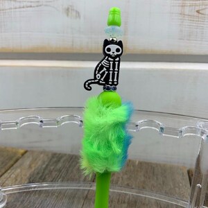 May include: A green pen with a fuzzy green and blue pom-pom top. The pen has a black skeleton cat charm with rhinestones.