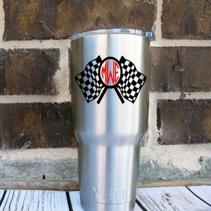 May include: A stainless steel tumbler with a checkered flag and monogram design. The tumbler has a black and white checkered flag with a red monogram in the center. The tumbler is on a white wooden surface.