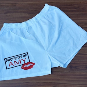 May include: White boxer shorts with an elastic waistband. The shorts feature the text "PROPERTY OF AMY" in a black rectangle, along with a red lip print.