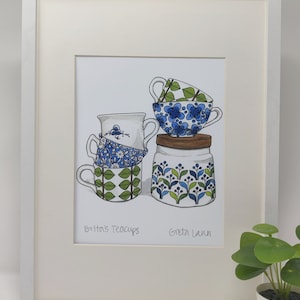May include: A framed illustration of four teacups and a jar with blue and green floral patterns. The text "Brita's Teacups" and "Greta Lann" are written below the illustration.