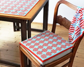 Könnte beinhalten: Ein Holztisch mit einer rot-weiß gemusterten Tischdecke. Zwei Stühle mit rot-weiß gemusterten Kissen stehen auf beiden Seiten des Tisches.