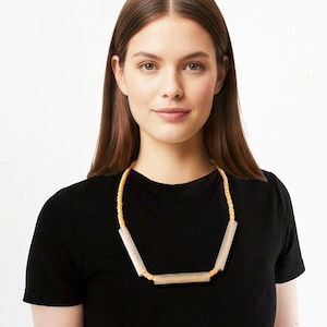 May include: A necklace featuring a light-colored, geometric pendant design. The pendant is connected by a braided, beige cord. The woman in the image is wearing a black t-shirt. The necklace is a modern, minimalist accessory.