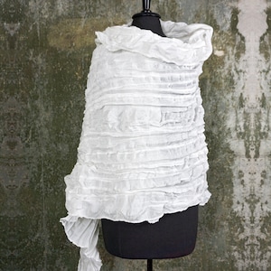 May include: White ruffled fabric shawl draped over a black mannequin. The shawl is made of a lightweight, sheer fabric with a crinkled texture.