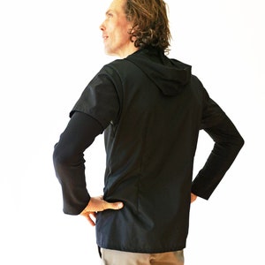 Puede incluir: Una camiseta negra de manga corta con capucha superpuesta sobre una capa base negra de manga larga. La prenda presenta una capucha en el cuello y una confecci&oacute;n en tejido negro liso y s&oacute;lido. Se lleva puesta con pantalones de color claro.