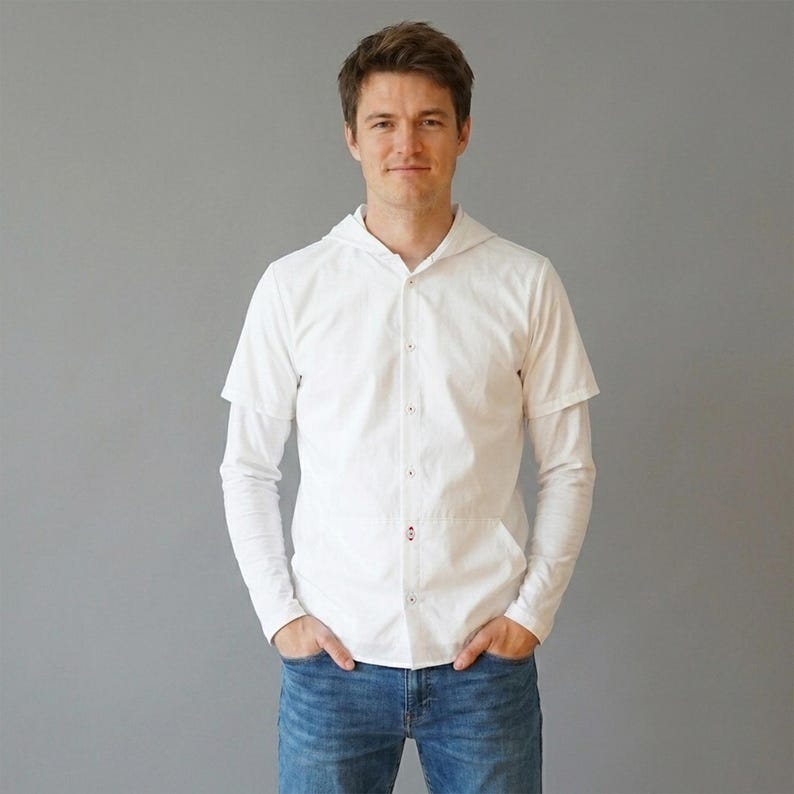 Puede incluir: Una camisa blanca de botones con capucha y mangas largas, que presenta un efecto de manga corta superpuesta en los brazos. La camisa tiene una tapeta de botones frontal y un peque&ntilde;o detalle rojo cerca del dobladillo, junto a unos vaqueros azules.
