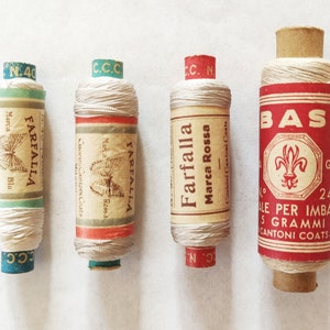 May include: Four spools of white thread with different colored labels. The labels have the text "Farfalla" and "Marca Rossa" on them. One spool has a label that says "Bast" with a fleur-de-lis design.