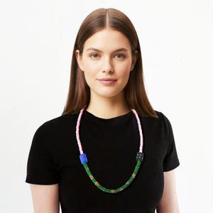 May include: A beaded necklace with a pink and white braided cord. The necklace features a green beaded section with red and yellow accents, and blue and black beaded square connectors.