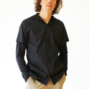 Puede incluir: Una camisa negra con botones, capucha y mangas cortas superpuestas sobre mangas largas. La prenda presenta una tapeta de botones frontal y una costura horizontal en la secci&oacute;n media, creando una apariencia de bolsillo. Tela de color negro liso.
