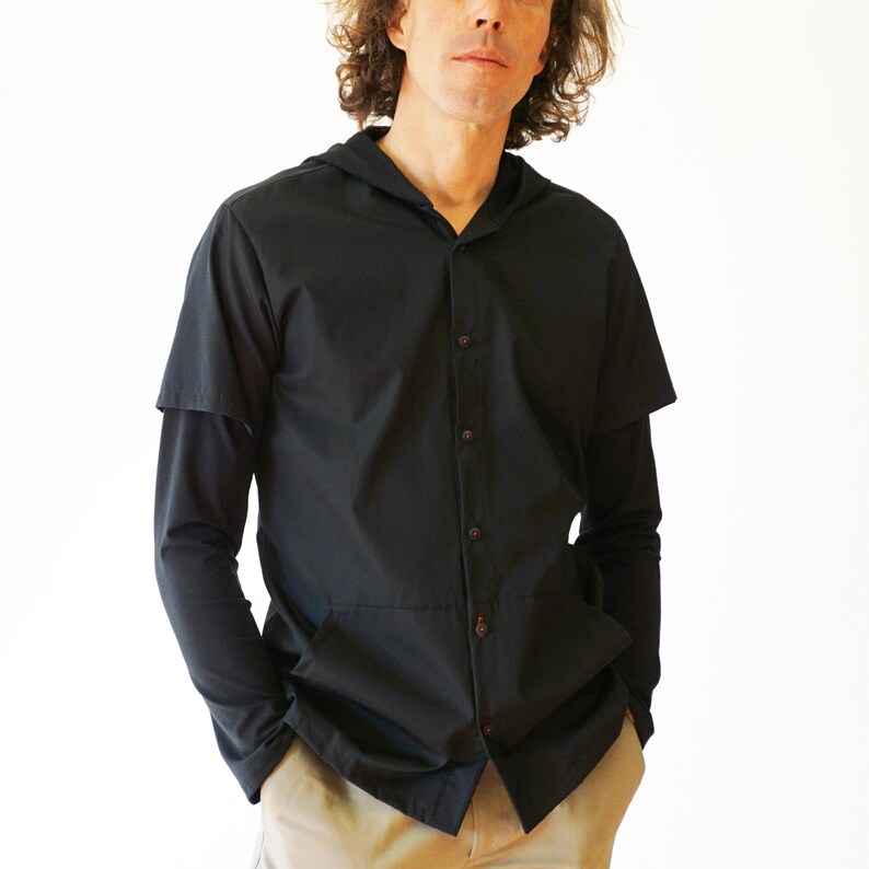 Puede incluir: Una camisa negra con botones, capucha y mangas cortas superpuestas sobre mangas largas. La prenda presenta una tapeta de botones frontal y una costura horizontal en la secci&oacute;n media, creando una apariencia de bolsillo. Tela de color negro liso.