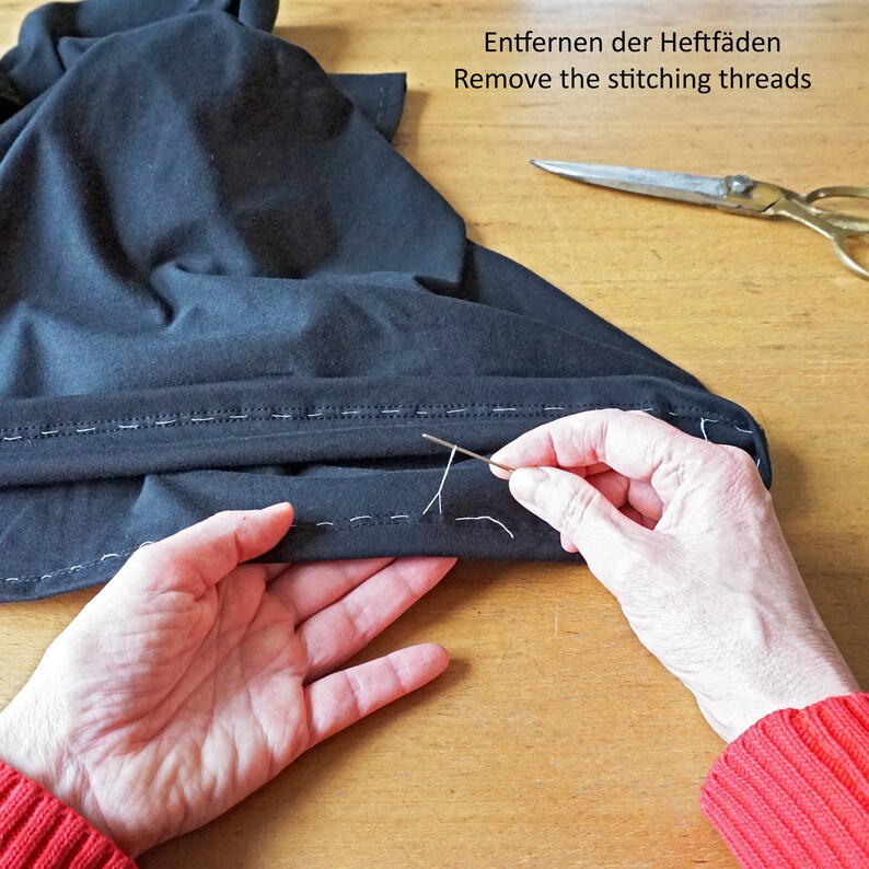 Puede incluir: Una prenda de tela negra con puntadas de hilv&aacute;n blancas a lo largo del dobladillo, una aguja de metal tirando de un hilo y unas tijeras de metal. El texto dice: Entfernen der Heftf&auml;den Remove the stitching threads.