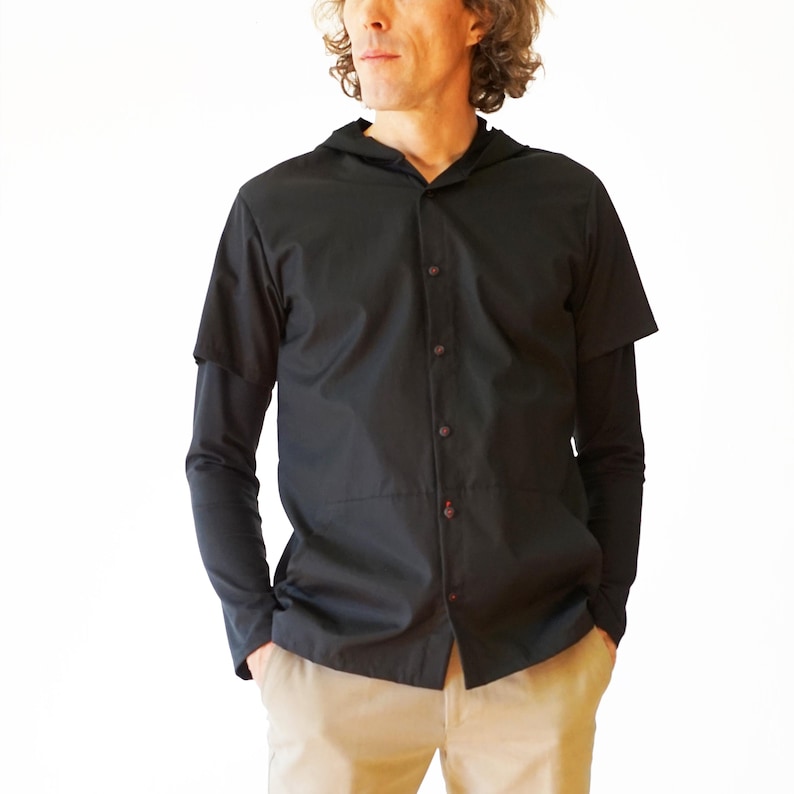 Puede incluir: Una camisa negra con capucha que presenta mangas cortas sobrepuestas a mangas largas, un cierre frontal de botones oscuros y una costura horizontal en la secci&oacute;n media. La prenda se combina con pantalones de color beige claro.