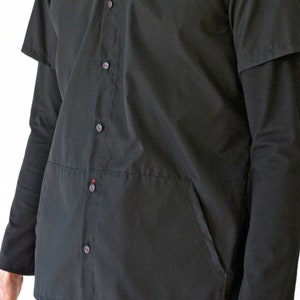 Puede incluir: Una camisa negra abotonada con mangas largas, una capa superpuesta de manga corta y un bolsillo frontal. La prenda incluye una tapeta vertical central con peque&ntilde;os botones negros, cada uno con un sutil detalle de hilo rojo en el centro.