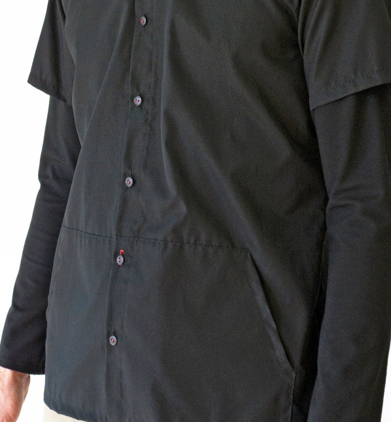 Puede incluir: Una camisa negra abotonada con mangas largas, una capa superpuesta de manga corta y un bolsillo frontal. La prenda incluye una tapeta vertical central con peque&ntilde;os botones negros, cada uno con un sutil detalle de hilo rojo en el centro.