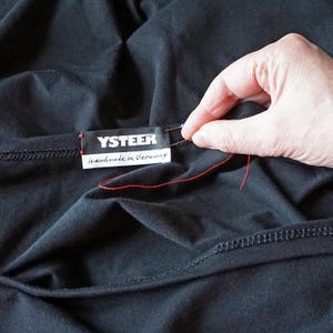 Puede incluir: Una prenda de tela negra con una etiqueta rectangular que muestra el texto blanco YSTEER y el texto m&aacute;s peque&ntilde;o Handmade in Germany. Se observa un hilo rojo unido a la etiqueta con una aguja, lo que indica el proceso de costura de la etiqueta.