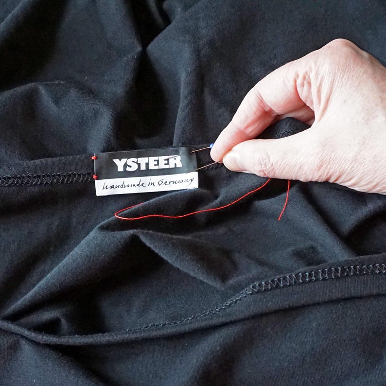 Puede incluir: Una prenda de tela negra con una etiqueta rectangular que muestra el texto blanco YSTEER y el texto m&aacute;s peque&ntilde;o Handmade in Germany. Se observa un hilo rojo unido a la etiqueta con una aguja, lo que indica el proceso de costura de la etiqueta.