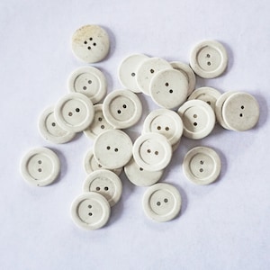 May include: A collection of white plastic buttons with two or four holes. The buttons are arranged in a random pattern on a white surface.