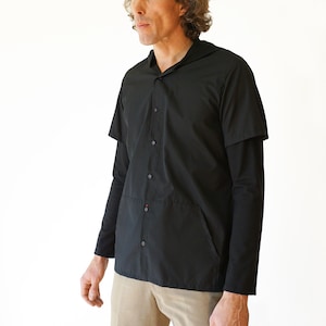 Puede incluir: Una camisa negra con botones, capucha y mangas cortas sobre mangas largas. La prenda presenta cierre frontal de botones, una costura horizontal en la secci&oacute;n media y bolsillos laterales integrados. Se combina con pantalones de color tostado.