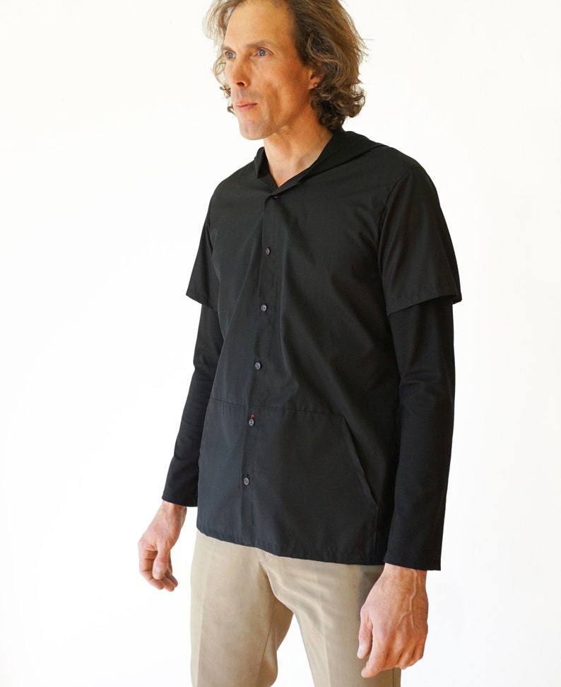 Puede incluir: Una camisa negra con botones, capucha y mangas cortas sobre mangas largas. La prenda presenta cierre frontal de botones, una costura horizontal en la secci&oacute;n media y bolsillos laterales integrados. Se combina con pantalones de color tostado.