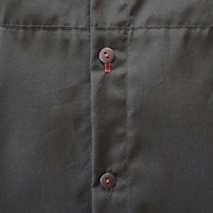Puede incluir: Una camisa negra con tapeta central que presenta dos botones circulares de color marr&oacute;n oscuro. Cada bot&oacute;n est&aacute; asegurado con hilo naranja brillante, que tambi&eacute;n resalta la costura vertical de los ojales sobre la tela negra lisa.