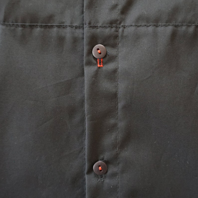 Puede incluir: Una camisa negra con tapeta central que presenta dos botones circulares de color marr&oacute;n oscuro. Cada bot&oacute;n est&aacute; asegurado con hilo naranja brillante, que tambi&eacute;n resalta la costura vertical de los ojales sobre la tela negra lisa.