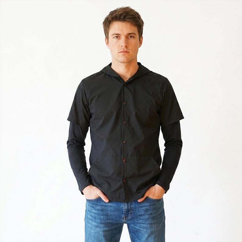 Puede incluir: Una camisa negra con capucha que presenta mangas cortas sobrepuestas a mangas largas, cierre frontal con botones y capucha. La prenda se combina con pantalones vaqueros azules. La camisa tiene un cuello estructurado y un acabado en negro liso.