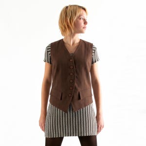 Op de afbeelding: Een bruin mouwloos vest met een V-hals, zes knopen aan de voorzijde en twee klepzakken. Het vest wordt gedragen over een jurk met korte mouwen en een zwart-wit zigzagpatroon. De outfit wordt gecompleteerd door een ondoorzichtige, donkerbruine panty.