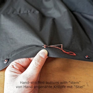 Puede incluir: Una prenda de tela negra con botones oscuros sujetos con hilo rojo vibrante. Una aguja de coser plateada atraviesa un bot&oacute;n, asegurado con un tallo de hilo. El texto "Hand-stitched buttons with "stem"" es visible en la imagen.
