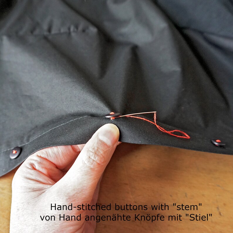Puede incluir: Una prenda de tela negra con botones oscuros sujetos con hilo rojo vibrante. Una aguja de coser plateada atraviesa un bot&oacute;n, asegurado con un tallo de hilo. El texto "Hand-stitched buttons with "stem"" es visible en la imagen.