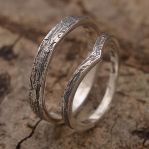 Könnte beinhalten: Zwei silberne Ringe mit strukturierten, rindenartigen Details. Ein Ring ist ein einfaches Band, während der andere ein V-förmiges Design hat. Die Ringe sind vor einem verschwommenen, hölzernen Hintergrund platziert und erzeugen eine rustikale Ästhetik.