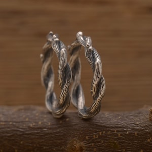 May include: Two silver rings with a twisted vine design. The rings are placed on a brown wooden branch, with a blurred wooden background. The rings have a textured surface, resembling tree bark.