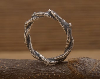 May include: A silver-toned ring with a twisted, branch-like design. The ring's band is formed by interwoven strands, creating a textured, natural appearance. The ring is displayed on a wooden surface, highlighting its organic aesthetic.