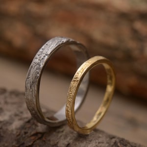 Puede incluir: Dos anillos de boda texturizados. Uno es plateado, el otro dorado. Las alianzas se colocan sobre un fondo de madera natural, resaltando su textura y diseño orgánicos. Los anillos probablemente son de metales preciosos.