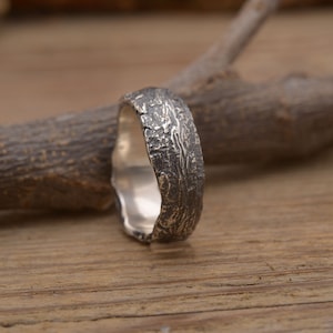 Op de afbeelding: Een zilveren ring met een getextureerd, op schors lijkend ontwerp. De ring heeft een brede band met een ruw, ongelijkmatig oppervlak, wat een rustieke esthetiek creëert. De ring wordt gepresenteerd op een houten oppervlak met een tak op de achtergrond.