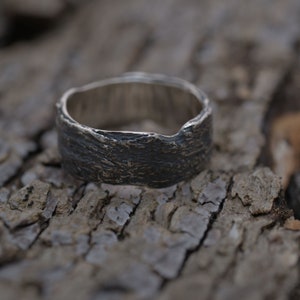 May include: A silver ring with a textured, bark-like design. The ring is thick and has a wide band.