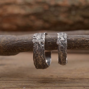 Puede incluir: Dos anillos de plata con un diseño texturizado que se asemeja a la corteza de un árbol. Los anillos están apilados uno encima del otro y descansan sobre una superficie de madera.
