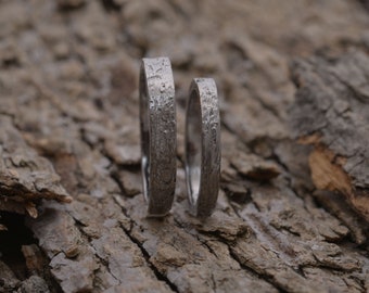 May include: Two silver wedding bands with a textured finish, resting on a piece of tree bark.