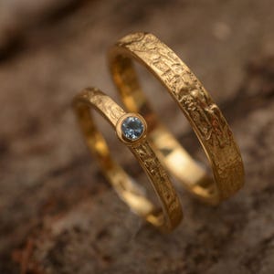 Puede incluir: Dos anillos de boda dorados. Un anillo presenta una pequeña gema redonda de color azul claro. Ambos anillos tienen un diseño texturizado y orgánico. Los anillos se muestran sobre un fondo neutro.
