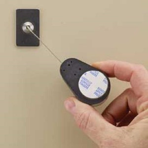 Puede incluir: Una etiqueta de seguridad negra con una pegatina adhesiva blanca que dice "Carefully Peel", unida a un cable delgado plateado. La etiqueta est&aacute; siendo sostenida por una mano.