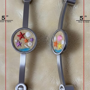 May include: Two silver-toned metal cabinet pulls with a 5-inch hole-to-hole measurement. Each pull features a circular inlay filled with sand, small colorful starfish, and seashells. The text SAMPLE ONLY and SOLD BLANK, NO GLASS INFUSED CABOCHONS is visible.
