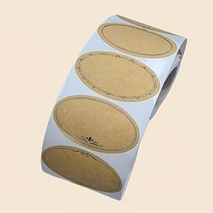 May include: A roll of oval-shaped kraft paper labels. Each label has a brown center with a decorative brown border and flourishes. The labels are on a white backing, ready to be peeled and used for various crafting or organizational purposes.