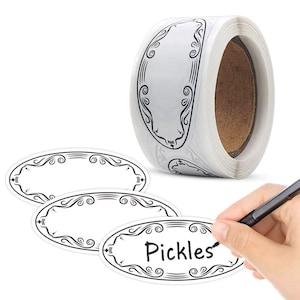 May include: A roll of white oval labels with black decorative borders. Three individual labels are shown, one with the word "Pickles" written on it. A hand is holding a pen, writing on a label.