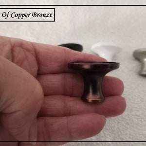 May include: A copper bronze cabinet knob is held in a hand, with other knobs in the background. The text "Hint Of Copper Bronze" is in the upper left corner. The knob has a dark brown finish with copper highlights.