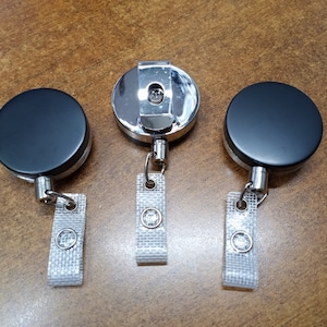 May include: Three black retractable badge reels with silver clips and clear straps. The reels are on a wooden surface.