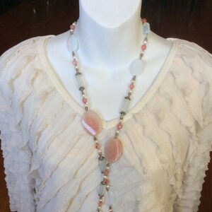 May include: A long necklace with pink and white oval-shaped beads and silver accents. The necklace is on a white ruffled shirt.