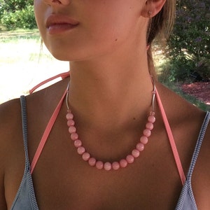 May include: A pink beaded necklace with a light pink ribbon and silver-colored metal accents. The necklace features round, light pink beads. The necklace is worn over a light blue top.