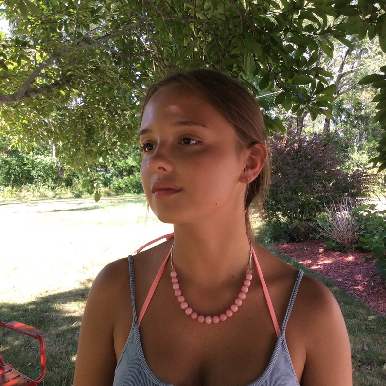 May include: A pink beaded necklace with a light pink ribbon and silver-colored metal accents. The necklace features round, light pink beads. The necklace is worn over a light blue top.