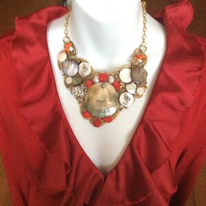 May include: A red ruffled blouse with a large, gold, and white seashell necklace. The necklace features a variety of shells, beads, and buttons.
